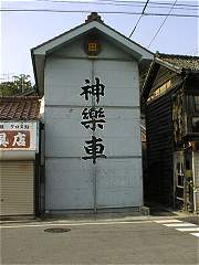 山車蔵・サヤ両側をアパート・民家に挟まれ、いかにも窮屈そうな倉．扉付きのシャッター．向かいは小学校屋根を解体して保管している